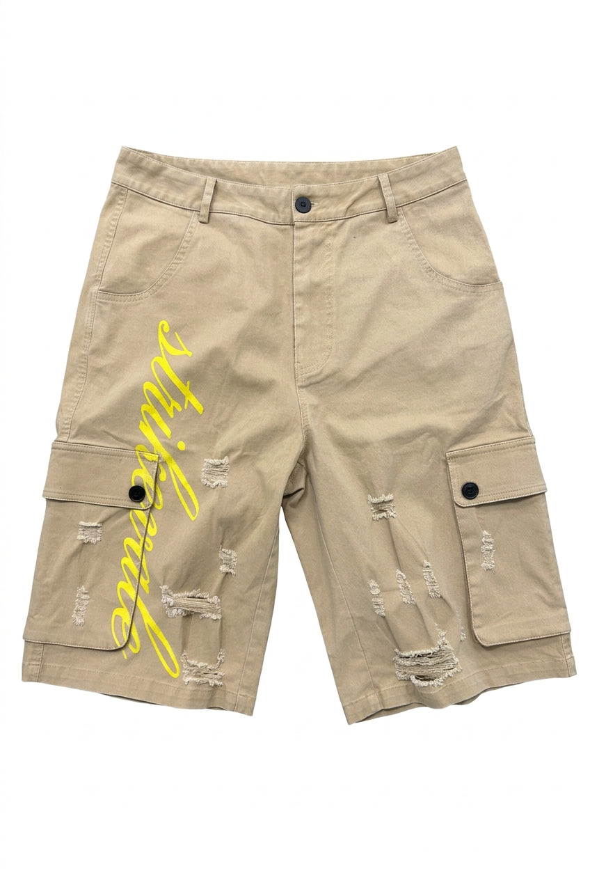 Beige cargo shorts with distressed rips and a yellow script graphic on the left leg, laid flat on a gray concrete floor with feet visible at the bottom.