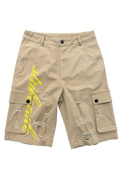 Beige cargo shorts with distressed rips and a yellow script graphic on the left leg, laid flat on a gray concrete floor with feet visible at the bottom.