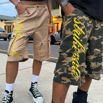 Two models wearing distressed cargo shorts with yellow script text. One model wears beige shorts and the other wears a green camouflage patterned shorts.