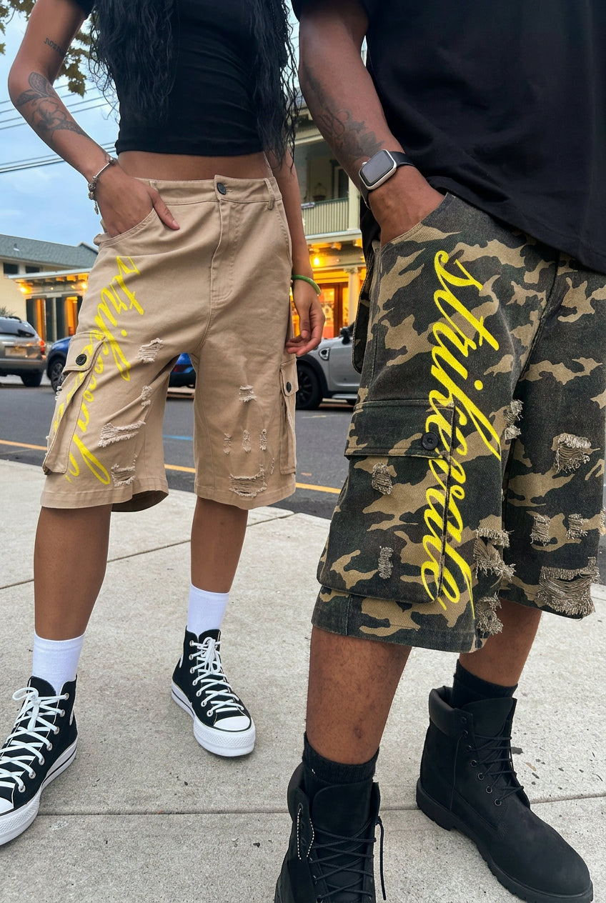 Two models wearing distressed cargo shorts with yellow script text. One model wears beige shorts and the other wears a green camouflage patterned shorts.