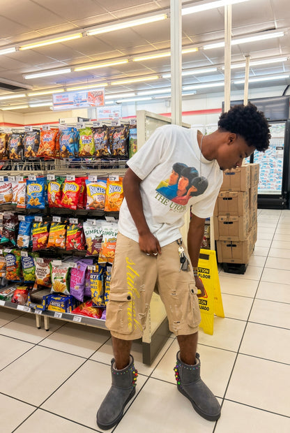 A man wearing a white graphic t-shirt, beige cargo shorts, and gray studded boots stands in a grocery store aisle looking down.